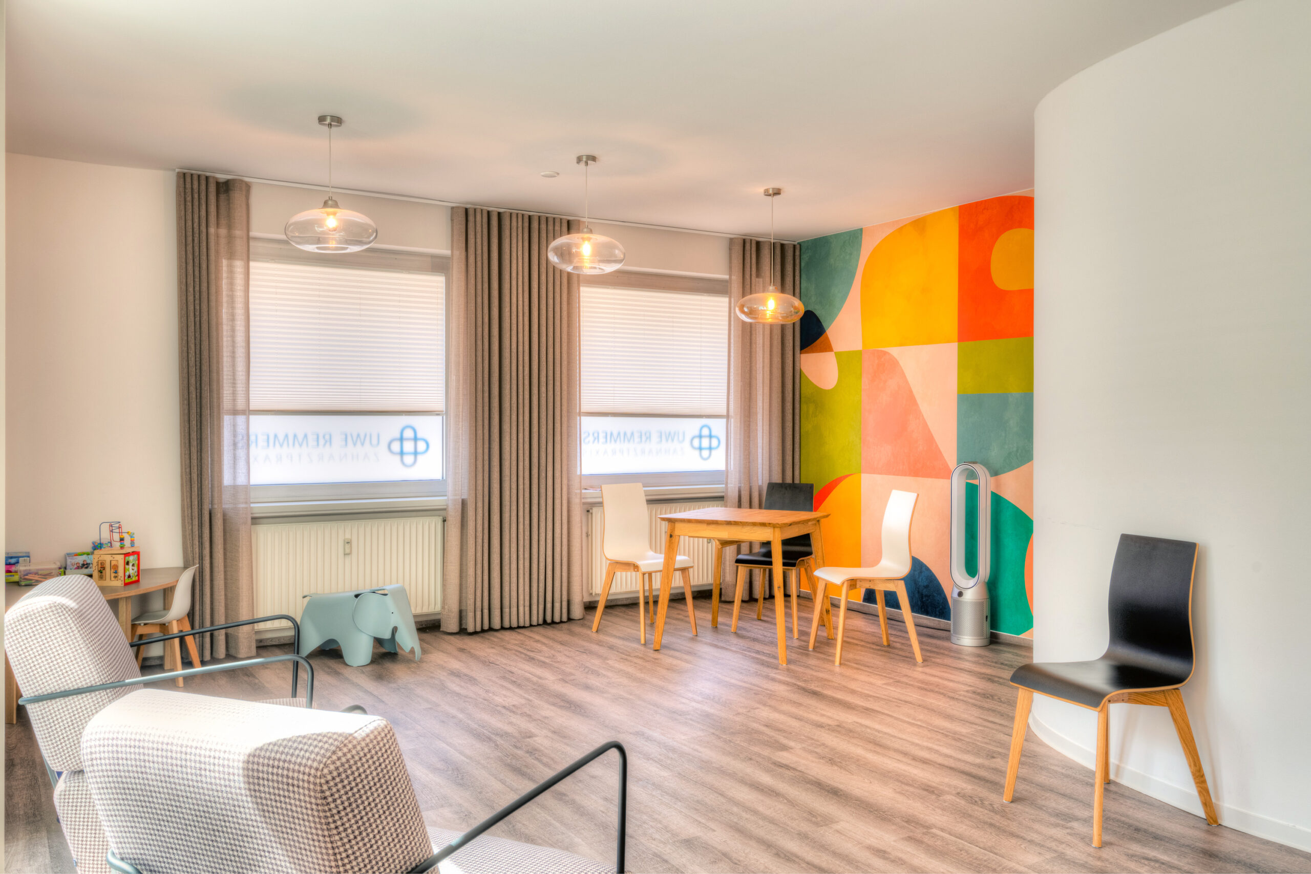 Praxis Remmers-20 Helle und moderne Wartezimmeratmosphäre in einer Zahnarztpraxis mit buntem Wanddesign, Holz- und Kunststoffmöbeln sowie Spielmöglichkeiten für Kinder.